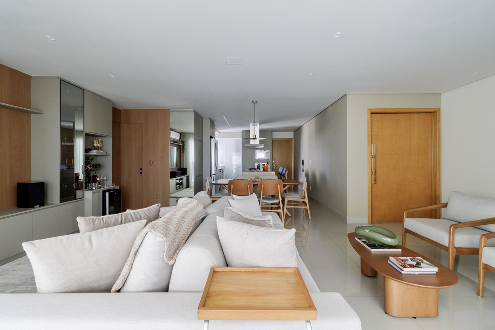 Sala de estar integrada com sofá claro, poltronas e mesa de centro orgânica em madeira, com a sala de jantar ao fundo, projeto do Anauá